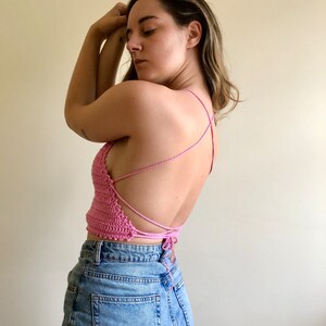 May include: A pink crocheted halter top with a criss-cross back design. The top is tied at the back and is worn with blue jeans.