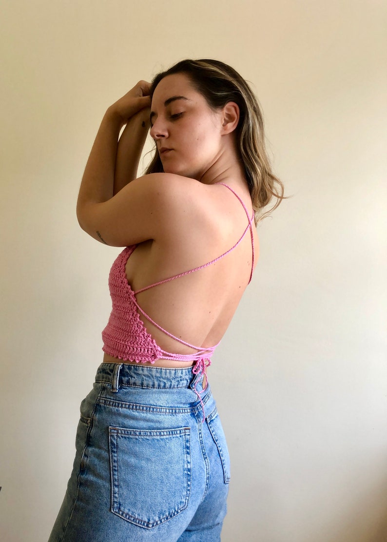 May include: A pink crocheted halter top with a criss-cross back design. The top is tied at the back and is worn with blue jeans.