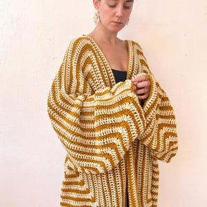 May include: A long, open-front crochet cardigan in a mustard yellow and cream striped pattern. The cardigan features wide sleeves and a geometric design on the lower portion. The garment is handmade and appears to be a comfortable, stylish layering piece.