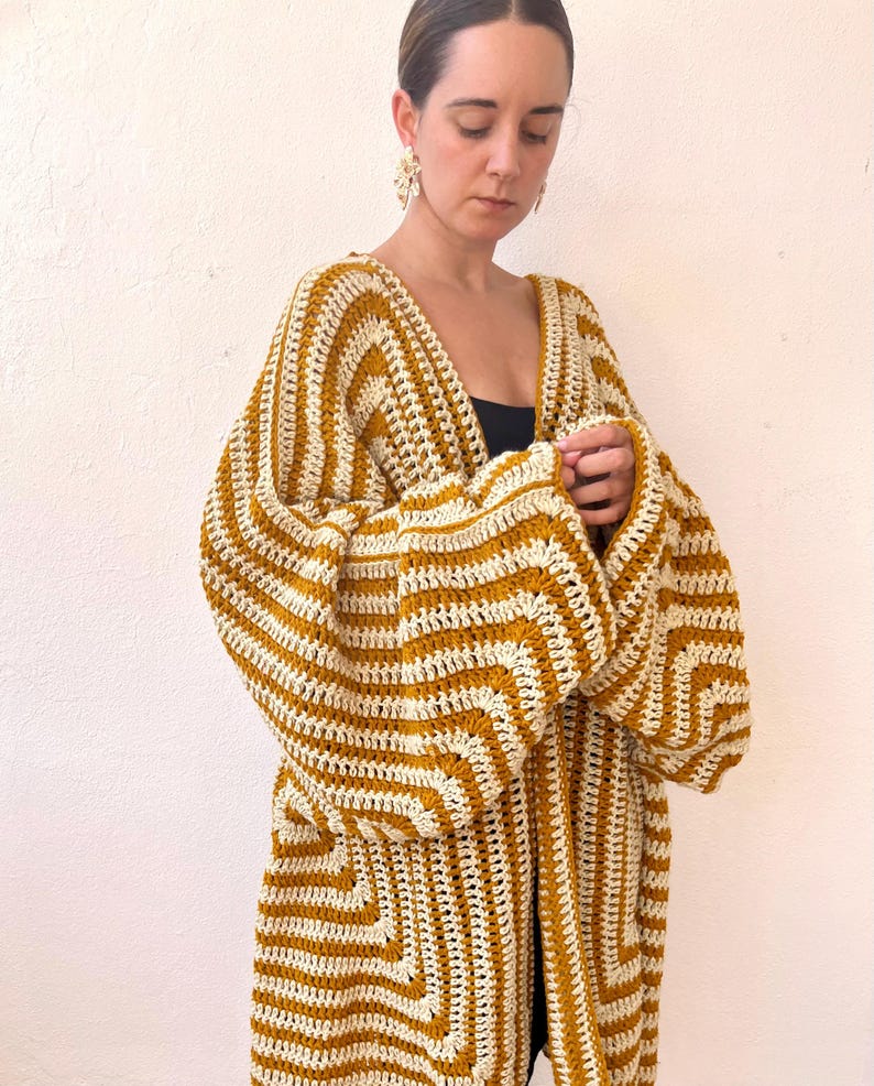 May include: A long, open-front crochet cardigan in a mustard yellow and cream striped pattern. The cardigan features wide sleeves and a geometric design on the lower portion. The garment is handmade and appears to be a comfortable, stylish layering piece.