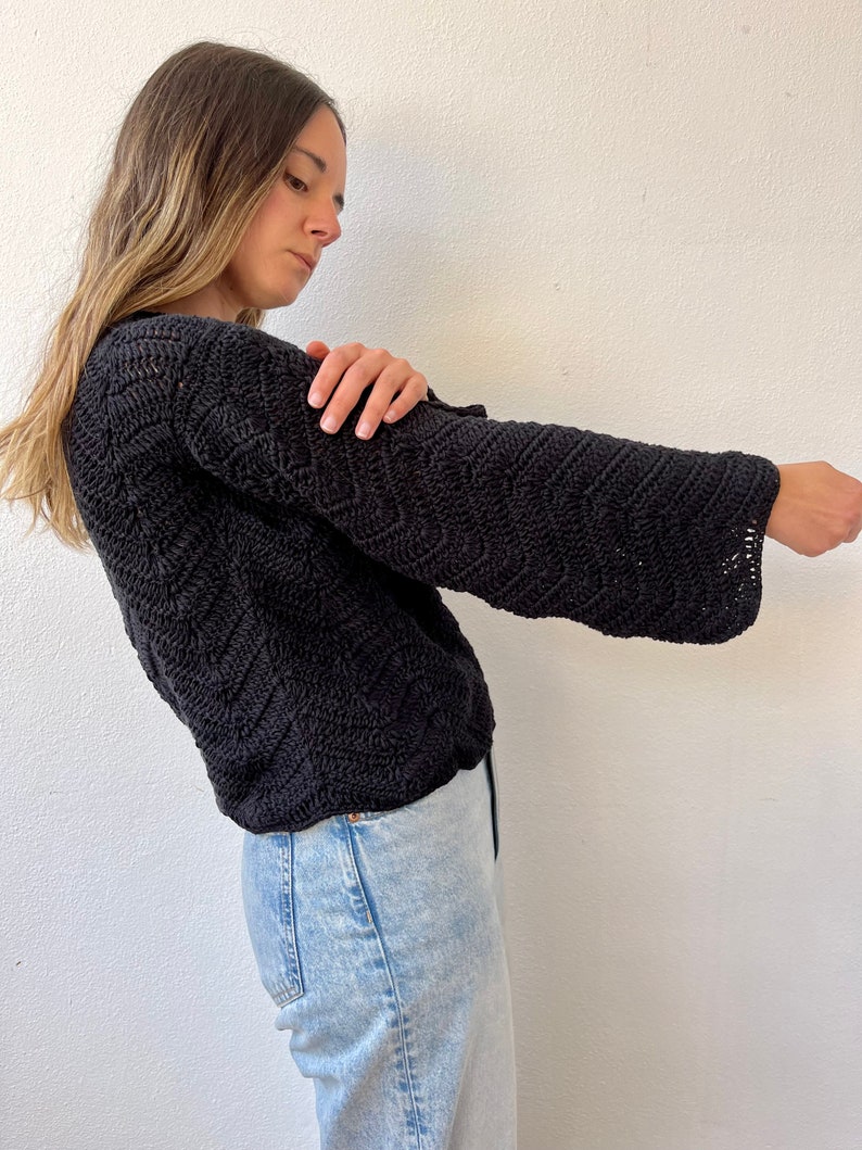 May include: A black crocheted sweater with a loose fit and long sleeves. The sweater has a textured pattern and is worn with light blue jeans.