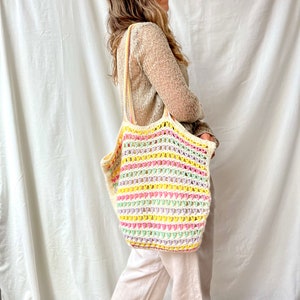 May include: A woman is carrying a white crochet tote bag with colorful stripes. The bag has a wide, open top and is made of a lightweight, breathable material.