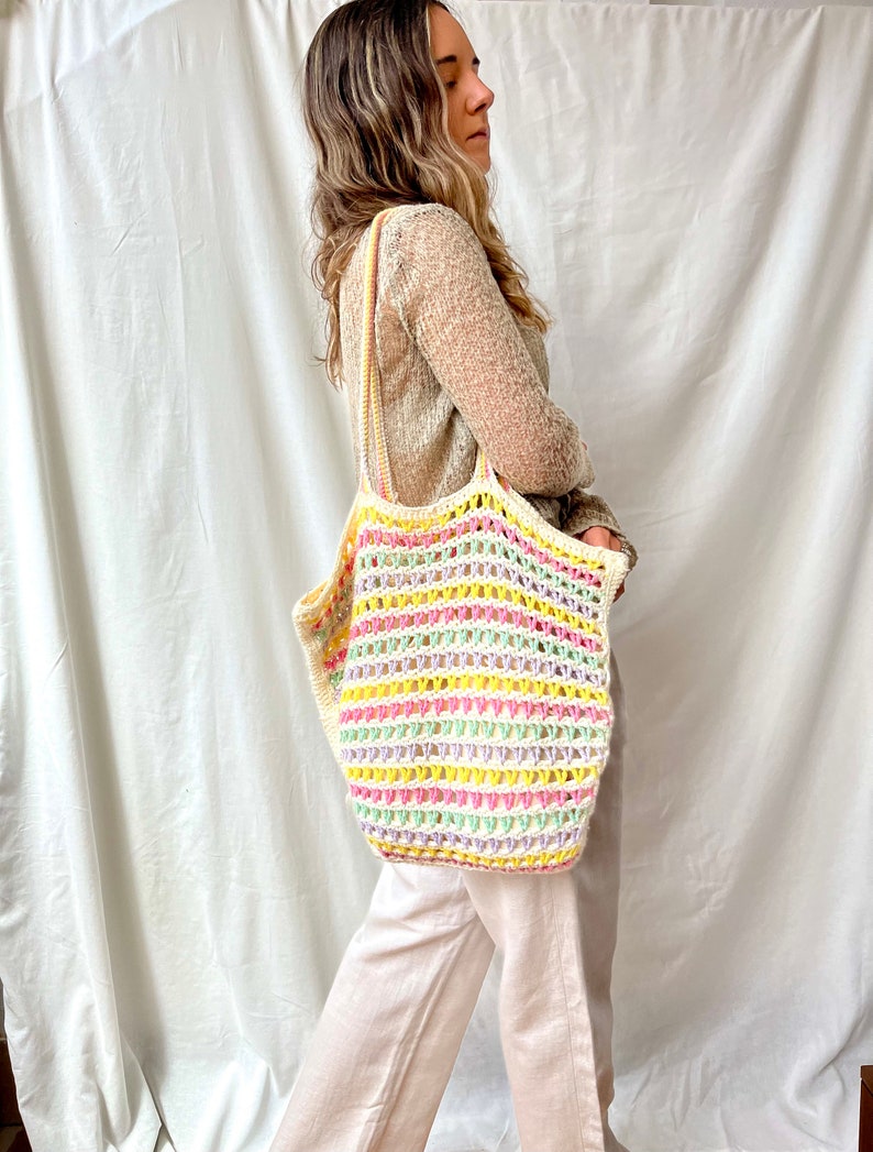 May include: A woman is carrying a white crochet tote bag with colorful stripes. The bag has a wide, open top and is made of a lightweight, breathable material.