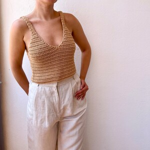 May include: A crocheted tank top in a light brown color. The top has a v-neck and thin straps. The model is wearing the top with white pants.
