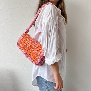 May include: A pink and orange crocheted shoulder bag with a long strap. The bag is worn by a person wearing a white shirt and blue jeans.