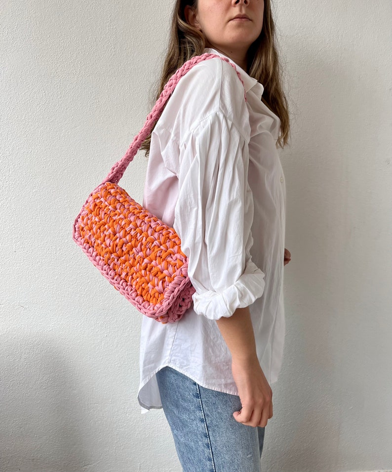 May include: A pink and orange crocheted shoulder bag with a long strap. The bag is worn by a person wearing a white shirt and blue jeans.