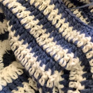 May include: Close-up of a handmade crochet item in a blue and white pattern. The texture of the yarn is visible, with a repeating design of alternating blue and white blocks. The item appears to be soft and cozy.