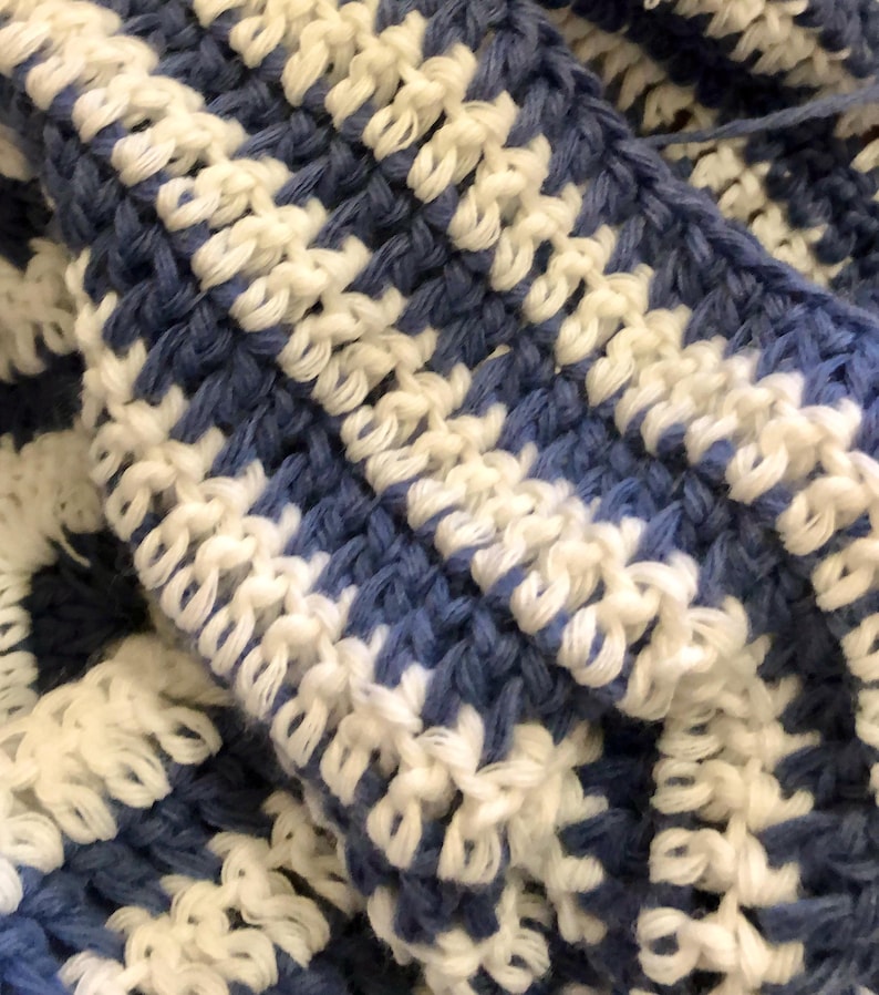 May include: Close-up of a handmade crochet item in a blue and white pattern. The texture of the yarn is visible, with a repeating design of alternating blue and white blocks. The item appears to be soft and cozy.