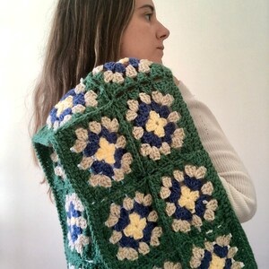 May include: A green, blue, and cream crochet blanket with a granny square pattern. The blanket is draped over a person's shoulders.