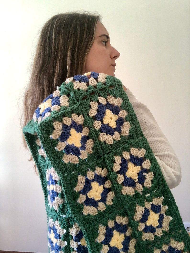May include: A green, blue, and cream crochet blanket with a granny square pattern. The blanket is draped over a person's shoulders.