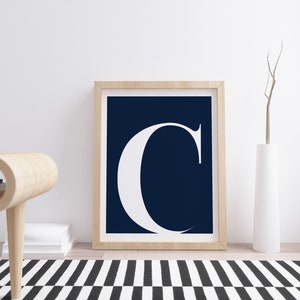 May include: A framed print with a white letter "C" on a dark blue background. The print is hanging on a white wall above a wooden table and a white vase with dried branches.