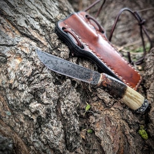 May include: A handcrafted knife with a textured blade and a handle made of bone and wood. The knife rests on a tree trunk next to a brown leather sheath with a leather strap.