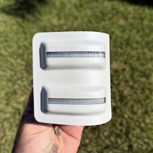 May include: A white plastic razor cartridge refill pack with two rectangular cartridges. Each cartridge has a blue and silver blade strip. The pack is held against a green grassy background.