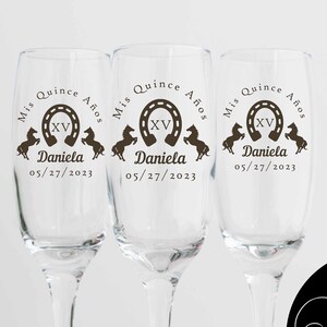 May include: Three clear champagne flutes with custom text. Each glass has the words "Mis Quince A&ntilde;os", a horseshoe with "XV", the name "Daniela", and the date "05/27/2023". The glasses are for a quincea&ntilde;era celebration.