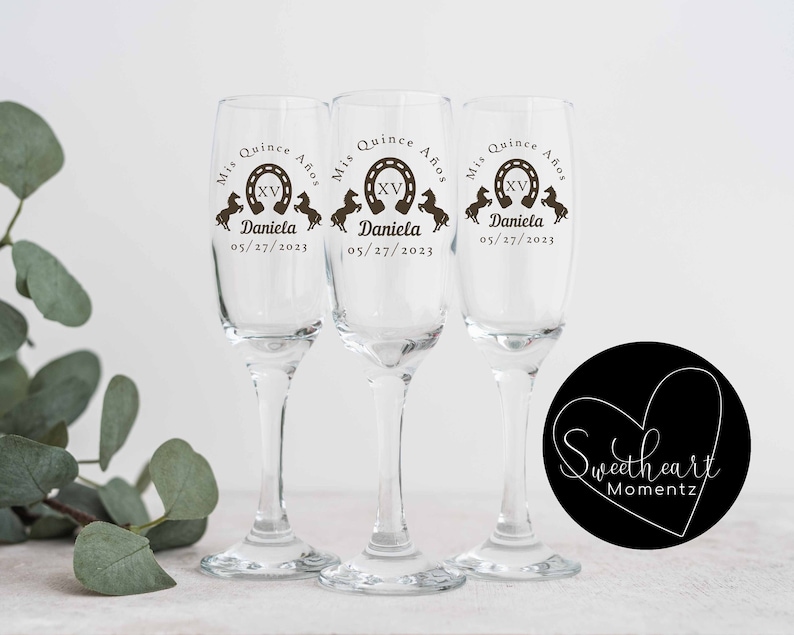 May include: Three clear champagne flutes with custom text. Each glass has the words "Mis Quince A&ntilde;os", a horseshoe with "XV", the name "Daniela", and the date "05/27/2023". The glasses are for a quincea&ntilde;era celebration.