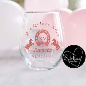May include: A clear stemless wine glass with a rose gold glitter design. The design includes the text "Mis Quince A&ntilde;os", a horseshoe, the Roman numeral XV, two horses, the name "Daniela", and the date 05/27/2023.