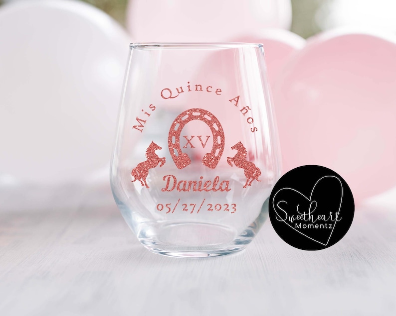 May include: A clear stemless wine glass with a rose gold glitter design. The design includes the text "Mis Quince A&ntilde;os", a horseshoe, the Roman numeral XV, two horses, the name "Daniela", and the date 05/27/2023.