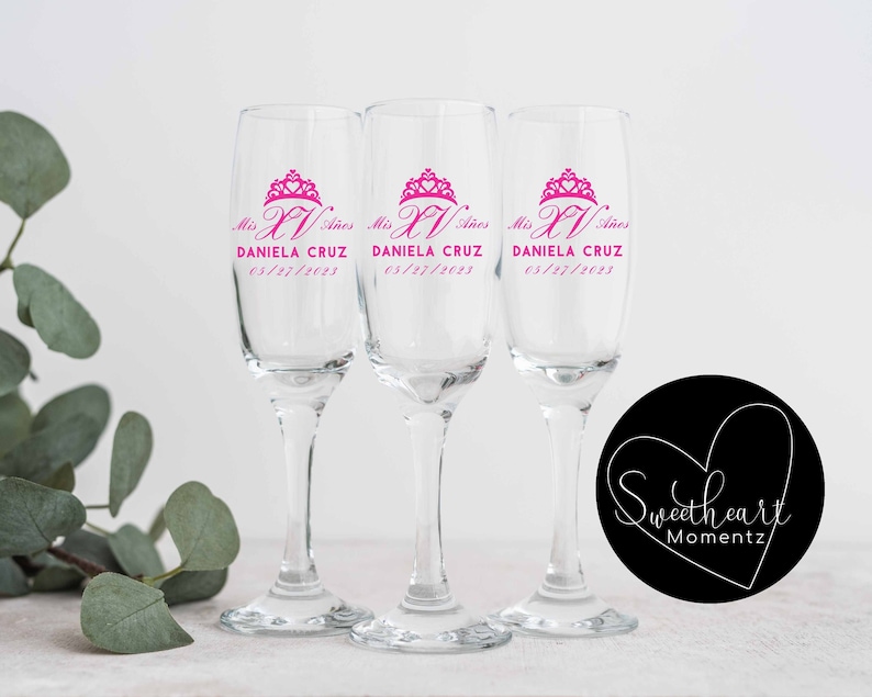 May include: Three clear glass champagne flutes with pink lettering that reads "Mis XV Anos" and "DANIELA CRUZ 05/27/2023".