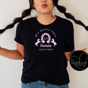 May include: Black t-shirt with a pink graphic design for a quincea&ntilde;era. The design includes the text "Mis Quince A&ntilde;os", a horseshoe, the Roman numeral XV, two horses, the name "Daniela", and the date "05/27/2023".