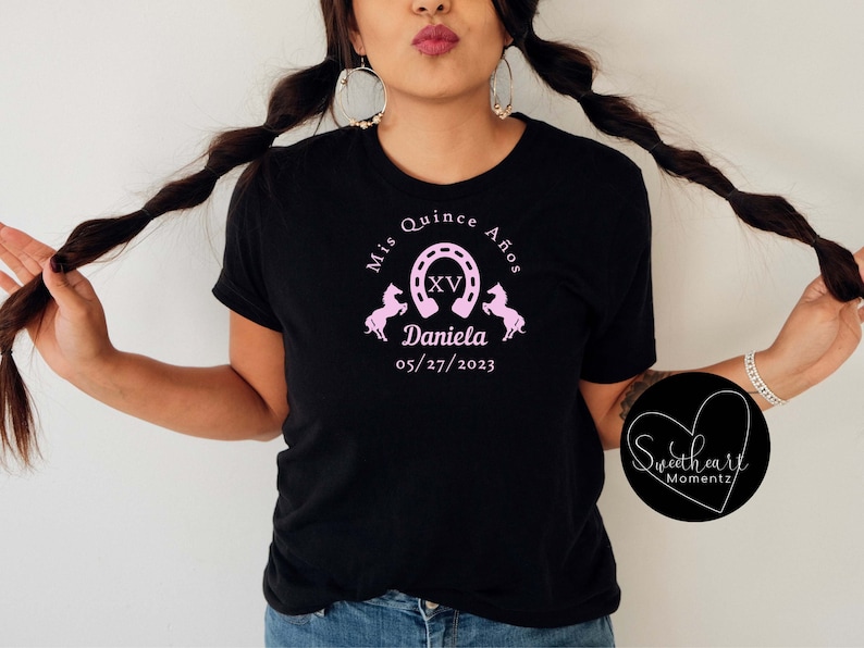 May include: Black t-shirt with a pink graphic design for a quincea&ntilde;era. The design includes the text "Mis Quince A&ntilde;os", a horseshoe, the Roman numeral XV, two horses, the name "Daniela", and the date "05/27/2023".