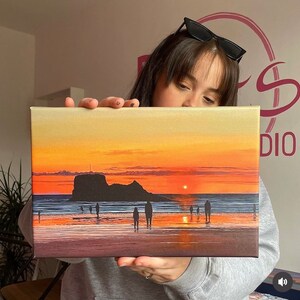 May include: A canvas painting depicting a sunset over a beach with a large rock formation in the distance. The sky is a vibrant orange and yellow, and the water is a calm blue. There are several small figures walking along the beach.