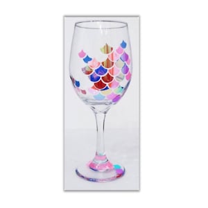 May include: A clear glass wine glass with a colorful mermaid scale design. The scales are painted in shades of pink, blue, purple, and green.