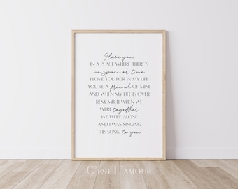 Op de afbeelding: Zwart-wit typografieprint met de tekst "I love you in a place where there's no space or time. I love you for in my life you're a friend of mine and when my life is over remember when we were together we were alone and I was singing this song to you." De print is ingelijst in een licht houten lijst en ligt op een lichte houten vloer. De tekst "C'EST L'AMOUR" is onder de lijst gedrukt.