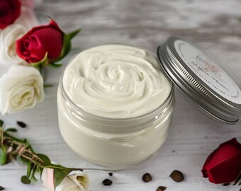 May include: A white jar of body butter with a silver lid. The jar is open and the body butter is swirled. The jar is surrounded by red and white roses and coffee beans.