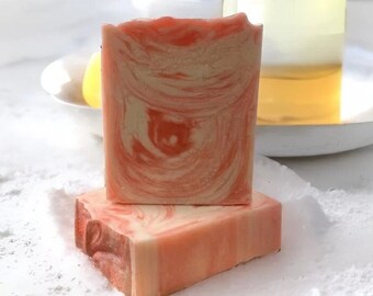 May include: Two rectangular bars of soap with a marbled pattern of coral orange and cream colors. One bar stands vertically while the other lies flat beneath it. Behind the soap is a clear glass bottle filled with a light yellow liquid.