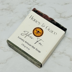 May include: A bar of soap with a black and green swirl pattern. The soap is wrapped in white paper with the text "Honey & Grace Silver Fox Luxury Artisan Bar Soap 170 g / 6 oz" printed on it.
