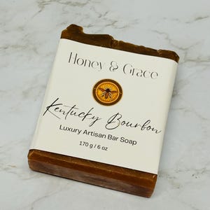 Puede incluir: Una barra de jabón marrón con una etiqueta blanca que dice "Honey & Grace Kentucky Bourbon Luxury Artisan Bar Soap 170g/6 oz". La etiqueta también presenta una ilustración de una abeja.