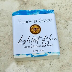 Op de afbeelding: Een zeepstaaf met een blauwe en witte swirl-ontwerp. Het label vermeldt "Honey & Grace" met een bijenlogo en "Lightest Blue" Luxury Artisan Bar Soap 170 g / 6 oz.