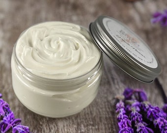 Può includere: Un barattolo di vetro pieno di burro corpo bianco e cremoso. Il barattolo ha un coperchio argentato con la scritta "Lavender Sweet" stampata su di esso. Il barattolo è appoggiato su una superficie di legno con fiori di lavanda viola.