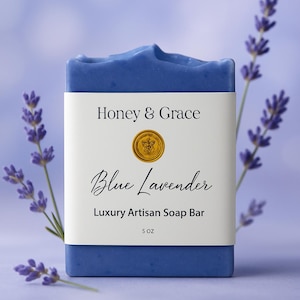 Puede incluir: Una pastilla de jabón artesanal de lavanda azul con una etiqueta blanca que dice "Honey & Grace Blue Lavender Luxury Artisan Soap Bar 5 oz." Ramas de lavanda enmarcan el jabón. El jabón pesa 142 gramos.