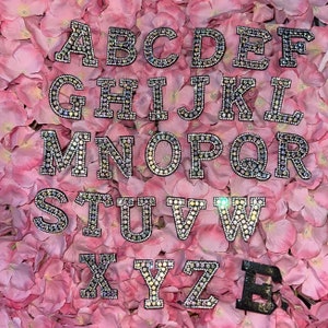 May include: A set of 26 alphabet letters made of black felt with rhinestones. The letters are arranged in alphabetical order and are displayed on a pink background.