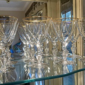 May include: A set of clear crystal wine glasses with gold rims. The glasses have a detailed, textured design on the bowl and a decorative stem. The glasses are displayed on a glass shelf, reflecting the light and enhancing their elegant appearance.