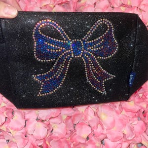 May include: Black cosmetic bag with a glittery finish, adorned with a large bow design made of colorful rhinestones. The bow features shades of blue, purple, and gold. The bag is set against a backdrop of pink flower petals.