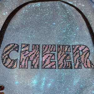May include: A light blue glitter backpack with the word "CHEER" in large, sparkling letters. The letters have a zebra print design with pink and black accents. The backpack has a black zipper and straps.