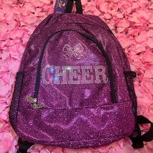May include: A sparkly purple backpack with the word "CHEER" spelled out in rhinestones. The backpack has a bow design above the word and a small charm that says "Bon Ange". The backpack has black straps and a mesh side pocket.