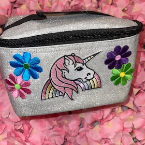 May include: Silver glitter cosmetic bag with a black zipper and handle. The bag features a unicorn patch with a pink mane and rainbow, and embroidered flower patches in blue, pink, purple, and green.