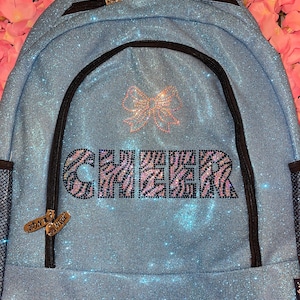May include: A light blue glitter backpack with black zippers and trim. The word "CHEER" is spelled out in a zebra print design. A pink bow is above the word "CHEER". Two small charms hang from the zipper.