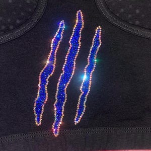 May include: Black top with three sparkling blue claw marks. The claw marks are outlined with iridescent rhinestones, creating a shimmering effect. The top has a scoop neckline and is made of a soft, dark fabric.
