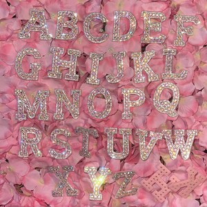 May include: A set of 26 alphabet letters made of rhinestones, each letter is individually crafted and features a sparkling design. The letters are arranged in alphabetical order and are displayed on a pink background.
