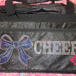 May include: Black glitter duffel bag with a large blue and purple rhinestone bow and the word "CHEER" spelled out in rhinestones. A pink dragon logo is on the lower right corner. The bag has a top zipper and two black handles.