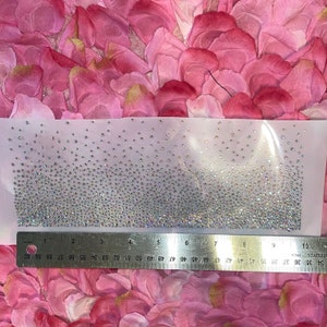 May include: A rectangular piece of clear fabric adorned with numerous small, sparkling rhinestones. The rhinestones are densely packed at the bottom, gradually thinning towards the top. A metal ruler with inch measurements is placed below the fabric.