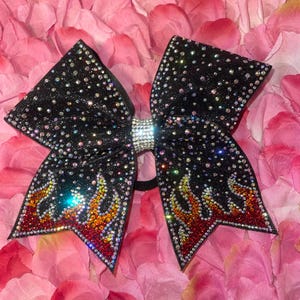 May include: Black cheerleading bow with a sparkling, glittery finish. The bow features a flame design in red, orange, and yellow rhinestones. A band of clear rhinestones accents the center of the bow, and the bow is set against a background of pink flower petals.