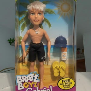Könnte beinhalten: Bratz Boyz Gabriel Puppe in einer Box. Die Puppe hat kurze, helle Haare, schwarze Shorts und eine Halskette. Zubehör umfasst eine blaue Kappe, Sonnencreme und gelbe Flip-Flops. Die Box zeigt eine Strandszene.