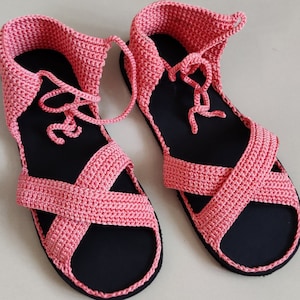 Op de afbeelding: Een paar roze gehaakte sandalen met zwarte zolen en veters. De sandalen hebben een kruislings ontwerp aan de bovenkant.