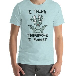 Puede incluir: Una camiseta azul claro con un drag&oacute;n de dibujos animados y el texto "Pienso, luego olvido".