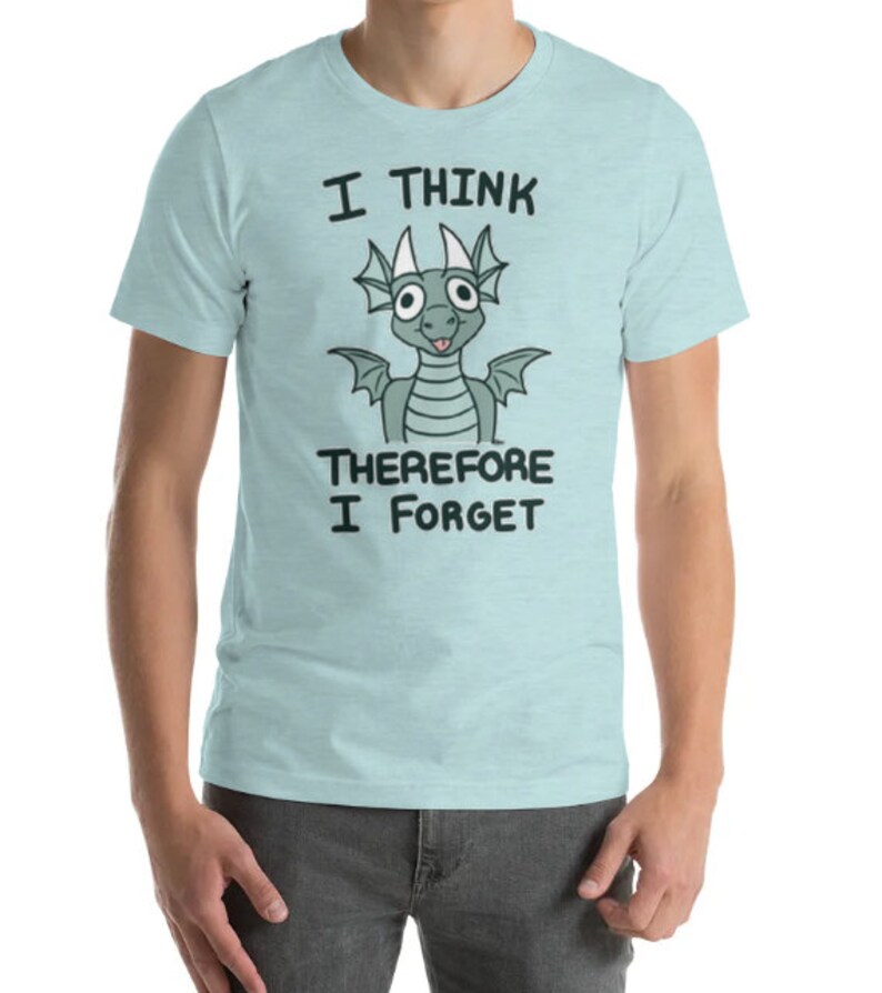 Puede incluir: Una camiseta azul claro con un drag&oacute;n de dibujos animados y el texto "Pienso, luego olvido".